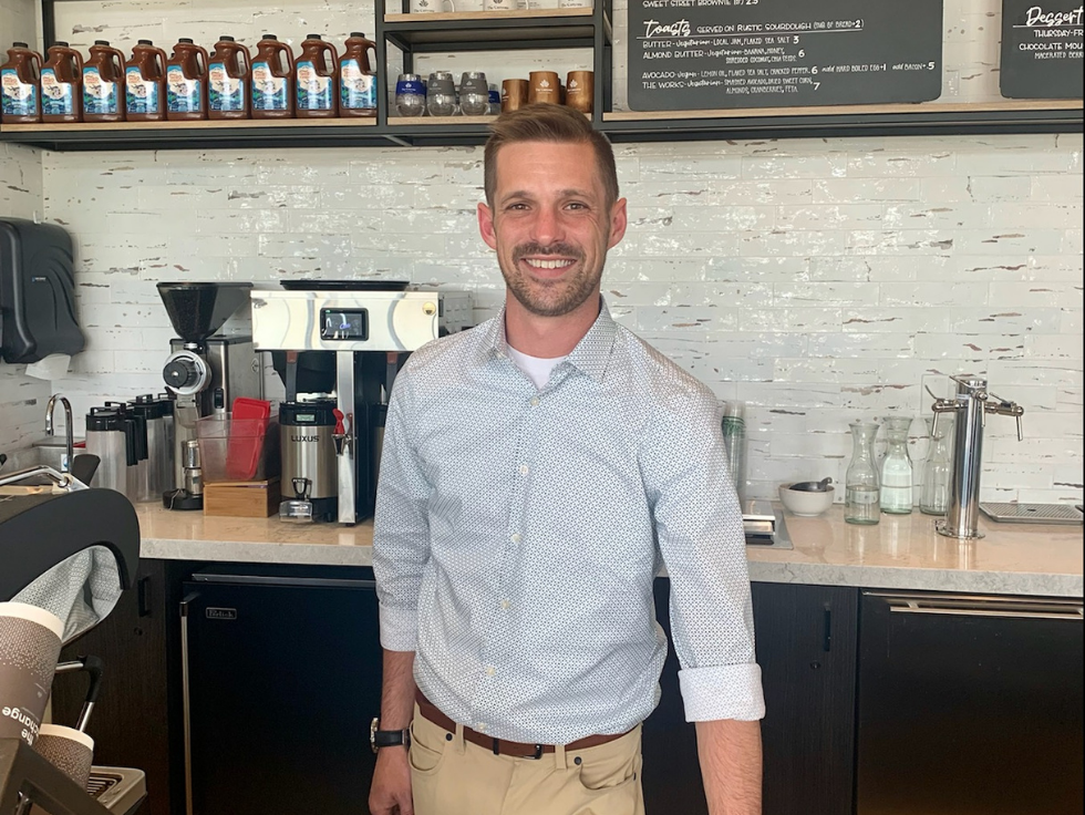 A man in a light blue shirt stands behind a coffee shop counter, smiling warmly. Shelves with coffee supplies and a menu board create a cozy backdrop, reminiscent of the inviting atmosphere you'd find in Castle Pines, known for its welcoming new home community and resort-style amenities.