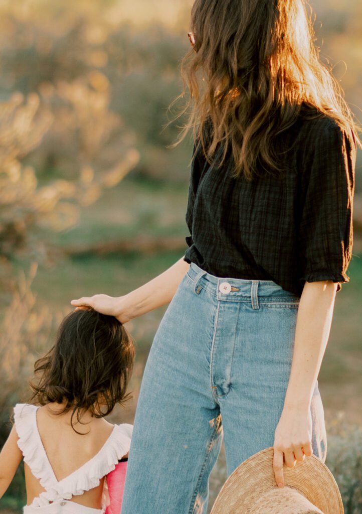 Woman in a black shirt and jeans stands outdoors, resting her hand on the head of a young girl in a white ruffled dress; the woman holds a straw hat, enjoying a sunny day near The Canyons new homes in Castle Pines.