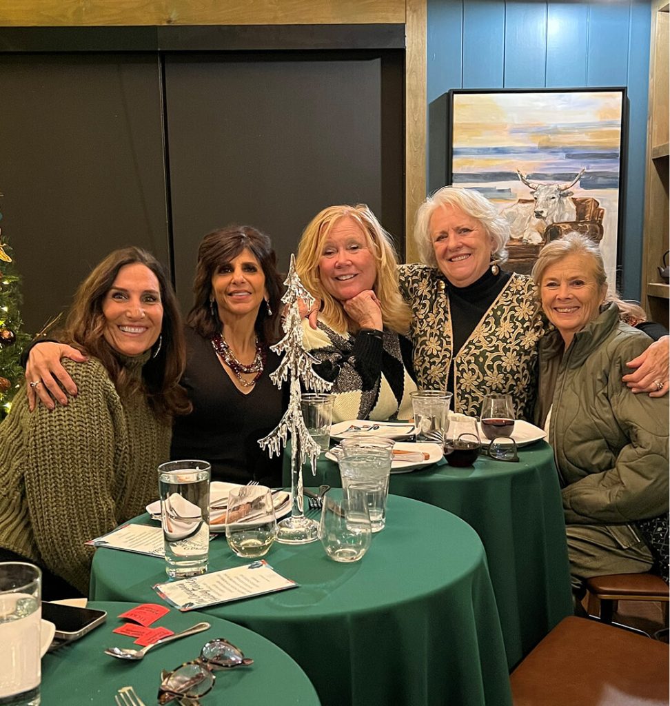 Five women sit around a table with green tablecloths in a restaurant, smiling at the camera. Place settings, glasses, and a menu are visible. A painting hangs on the wall behind them.